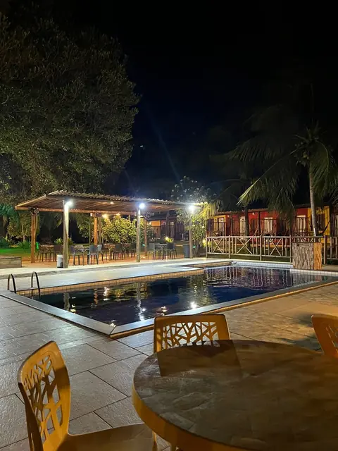 Pérgola com iluminação suave, mesas ao redor e piscina refletindo as luzes da pousada.